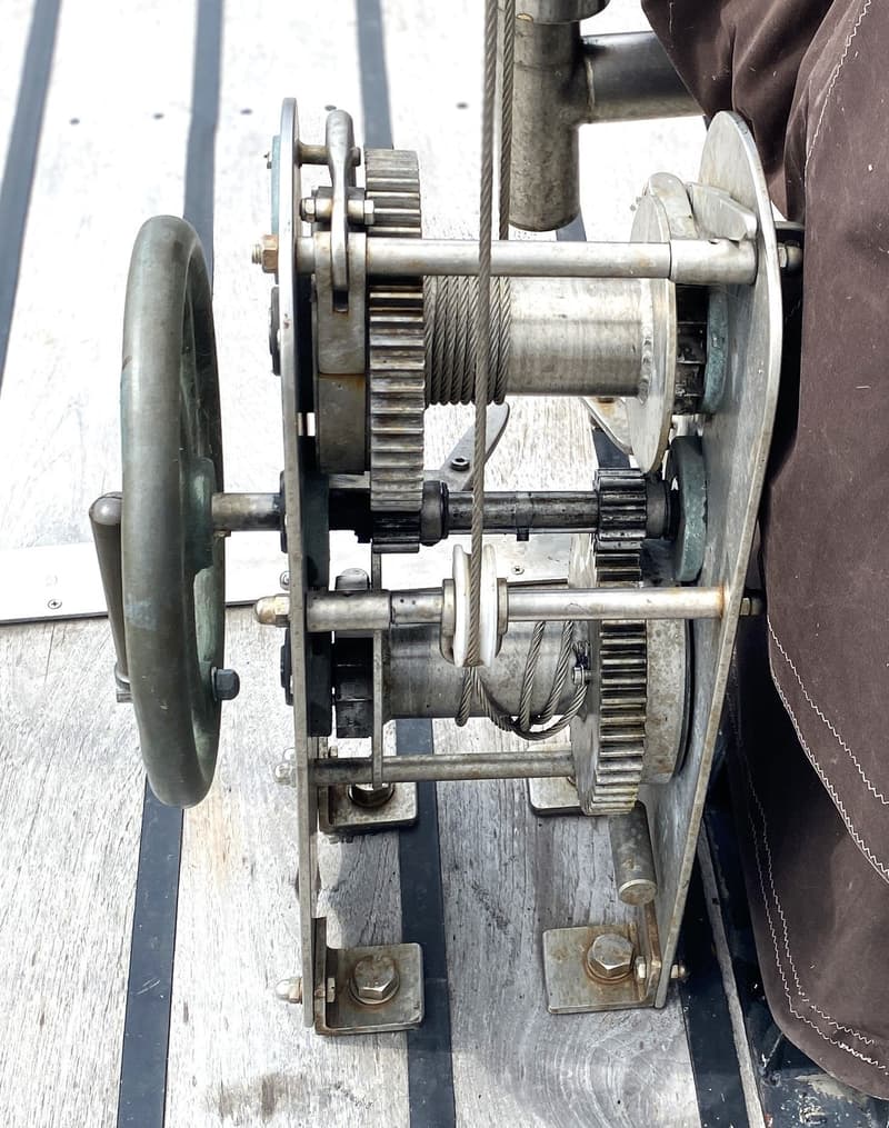 Een detailfoto van een metalen ankerlier met tandwielen en kabels, gemonteerd op een houten dek.