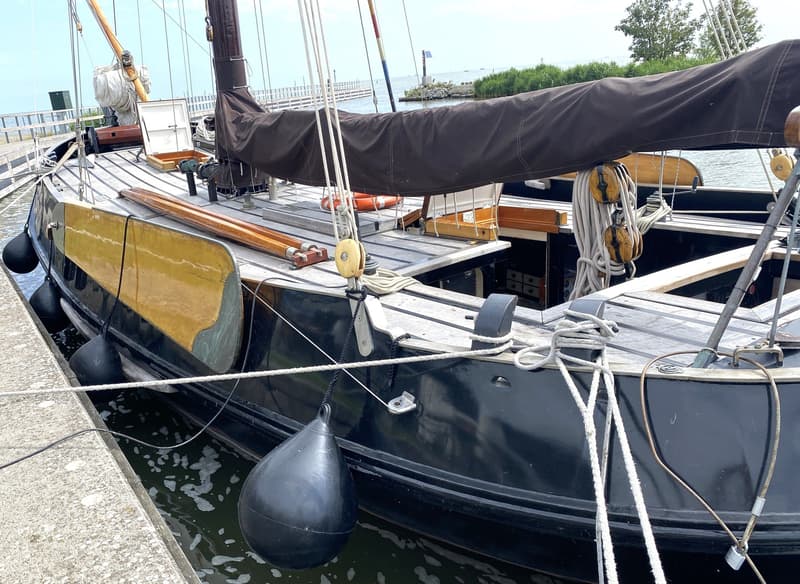 Een zeilboot met een zwart zeil en een houten dek ligt afgemeerd aan een betonnen kade. — Schokker 14.00 M. One Off