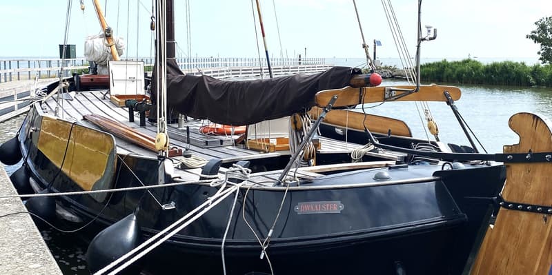 Een donkerblauwe boot met de naam "DWAALSTER" ligt afgemeerd aan een steiger, met een houten roer en een zwart zeil.