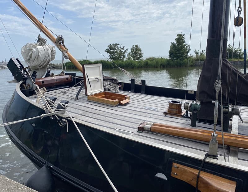 Een donkergekleurde boot met een houten dek en een open luik, afgemeerd aan een kade met water en groen op de achtergrond.