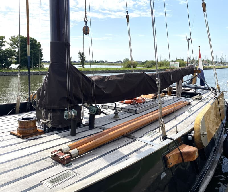 Een houten dek met een donkerbruin zeil, mast en diverse lijnen, met een Nederlandse vlag aan de achtersteven.