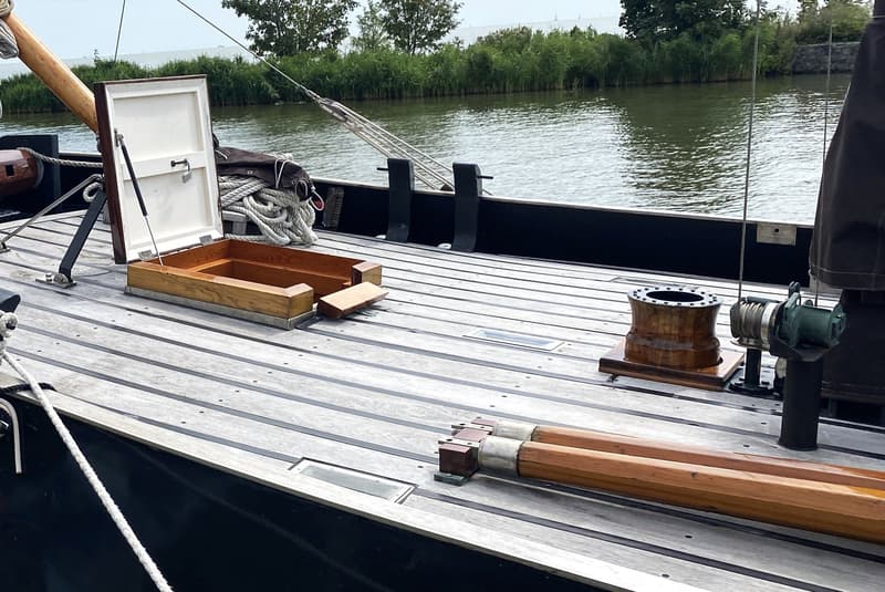 Een houten dek met een open luik, een lier, een kaapstander en twee houten masten, met water en groen op de achtergrond.