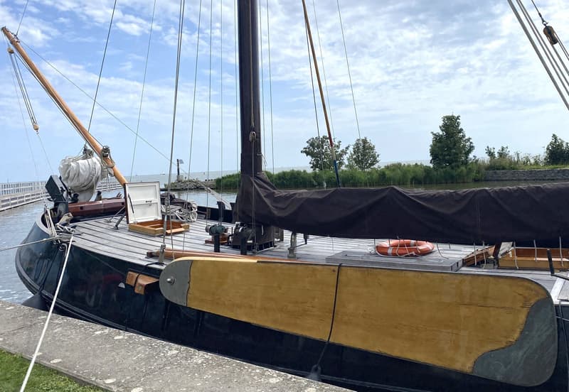 Een donkerblauwe zeilboot met een houten dek en een zwart zeil ligt afgemeerd aan een kade. — Schokker 14.00 M. One Off