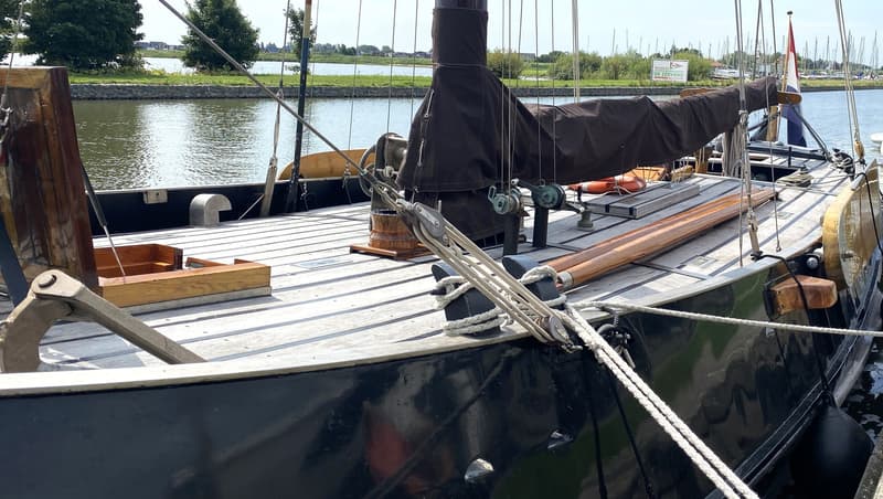 Een zwart zeiljacht met een bruin zeil en een Nederlandse vlag ligt aangemeerd aan een kade.
