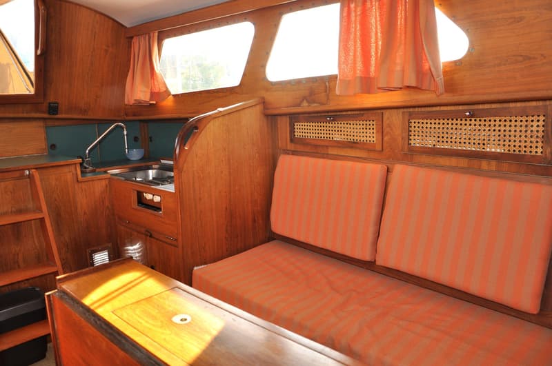 De foto toont een houten interieur van een boot met een kombuis, oranje gestreepte kussens en ramen met oranje gordijnen.