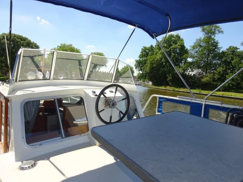 Een boot met een stuurwiel, voorruit, blauwe bimini en een tafel op het achterdek, varend op een kanaal.
