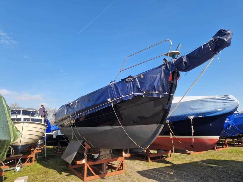Een donkerblauwe zeilboot met een blauw dekzeil staat op houten bokken op het gras onder een helderblauwe lucht.