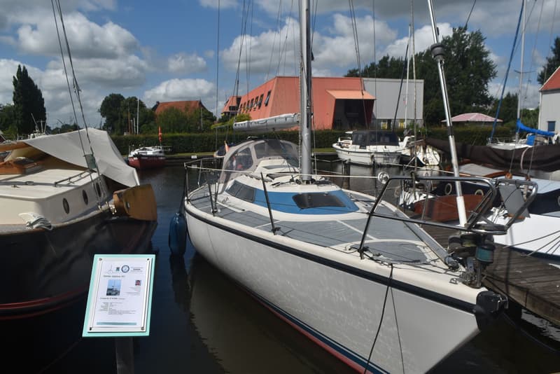 Een witte zeilboot met blauwe accenten ligt aangemeerd in een haven, met een advertentiebord ernaast. — Dehler Optima 101
