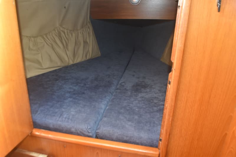 De foto toont een kajuit met twee blauwe matrassen, houten wanden en een beige stoffen opbergvak. — Dehler Optima 101