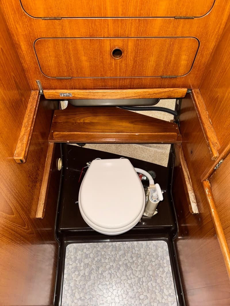 Een houten kajuit met een wit toilet, een donkerbruine vloer en een grijze vloermat met kiezelpatroon. — Dehler Optima 101