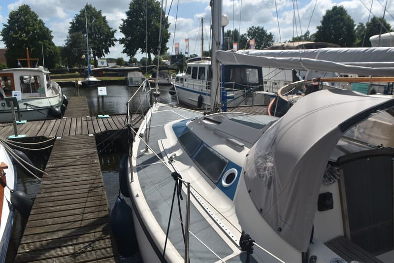 Een witte zeilboot met blauwe accenten en een grijze buiskap ligt afgemeerd aan een houten steiger. — Dehler Optima 101