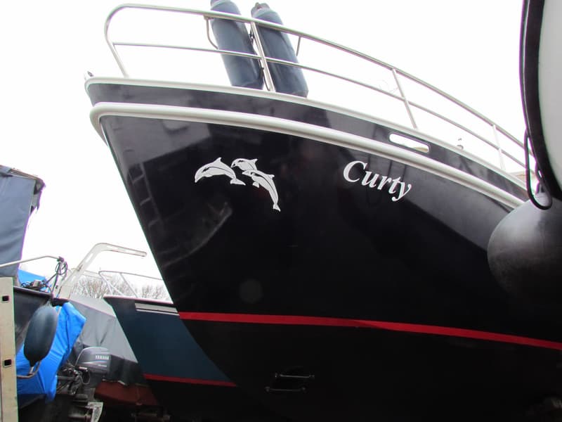 De boeg van een donkere boot met de naam "Curty" en een dolfijnmotief, met stootwillen en een reling. — Curtevenne 950 GSAK
