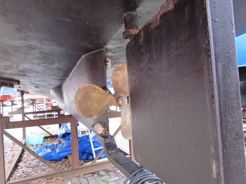 De foto toont de onderkant van een boot met een bronzen schroef en een roer, ondersteund door een stalen constructie.