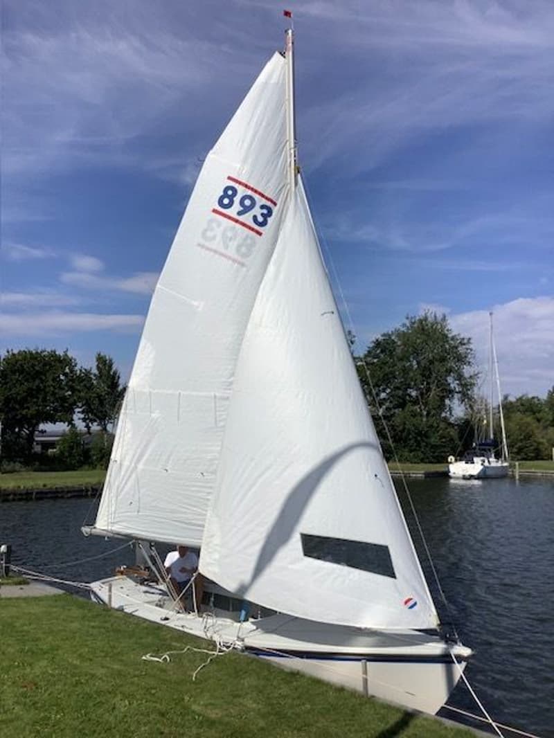 Een witte zeilboot met twee witte zeilen ligt aangemeerd, met "893" in blauw op het voorzeil en een persoon in de kuip.