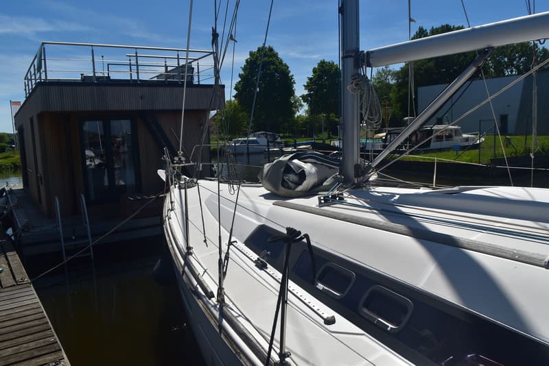 Een witte zeilboot ligt afgemeerd naast een houten woonboot, met een opgerold zeil op het dek. — Beneteau Oceanis 37