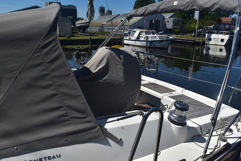 Een witte zeilboot met een grijze huik en stuurstandhoes ligt in een haven met andere boten en gebouwen op de achtergrond.