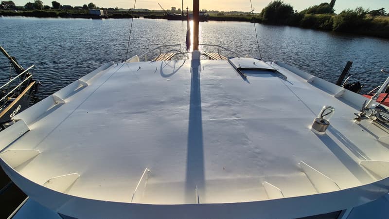 De foto toont het voordek van een boot met een mast en een luik, met water en landschap op de achtergrond. — Botter 14.99