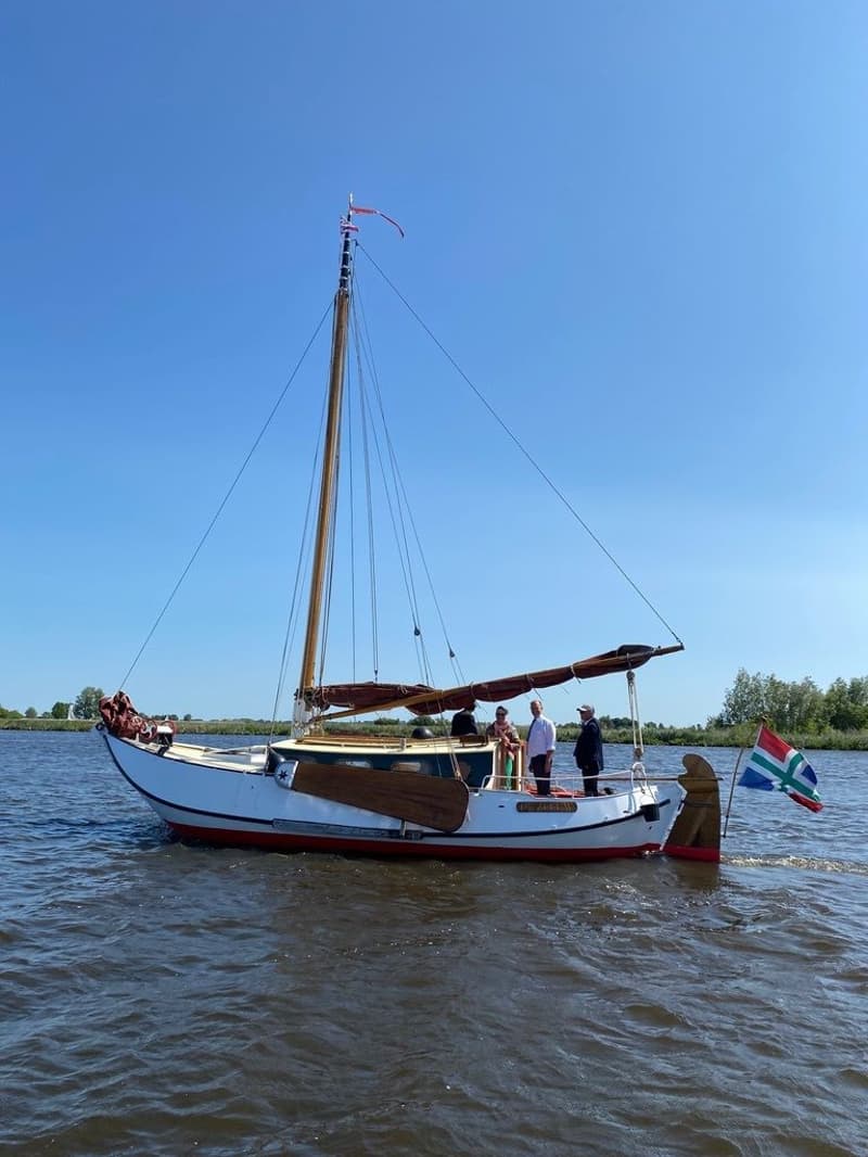 Een witte zeilboot met rode onderkant en houten details vaart op het water met vier personen aan boord.