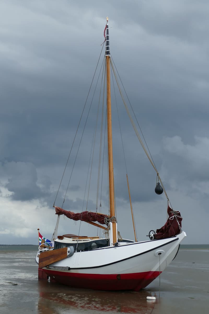 Een witte zeilboot met rode onderkant en een houten mast staat op een zandstrand onder een bewolkte lucht.