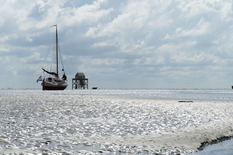 Een zeilboot met een Nederlandse vlag ligt op een zandbank bij laagwater, met een paalwoning en een andere boot op de…