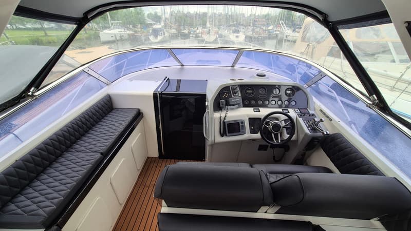 De foto toont de cockpit van een boot met een stuur, instrumentenpaneel, zwarte gestoffeerde banken en een houten vloer.
