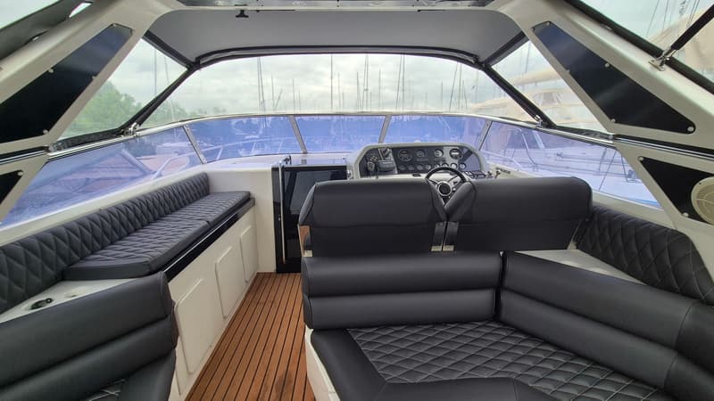 De foto toont het interieur van een boot met een stuurstand, zwarte gestoffeerde zitbanken en een houten vloer.