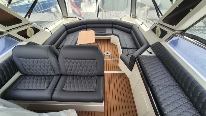 De foto toont het interieur van een boot met een U-vormige zwarte bank, een houten tafel en een houten vloer.