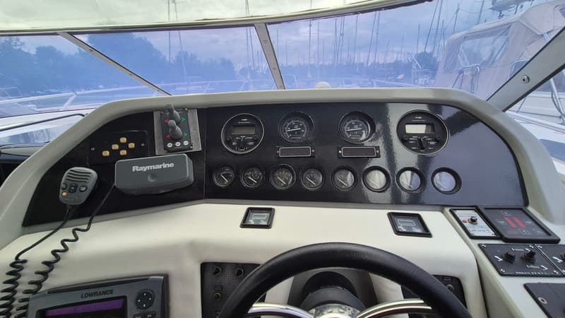 Een stuurstand met een stuurwiel, diverse meters, een Raymarine marifoon, een Lowrance dieptemeter en een uitzicht op een…