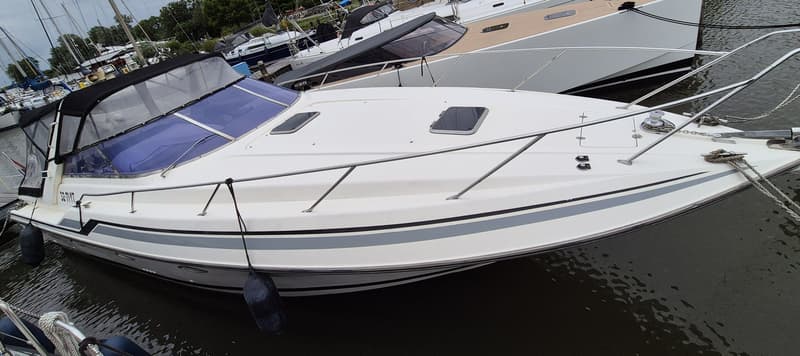 Een witte motorboot met een zwarte kap en blauwe ramen ligt in het water, met andere boten op de achtergrond.