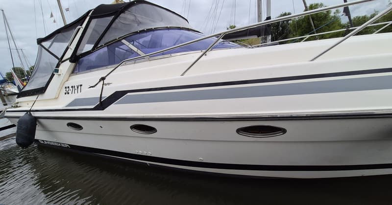 Een witte boot met een zwarte en grijze streep, patrijspoorten en een zwarte buiskap, ligt in het water.