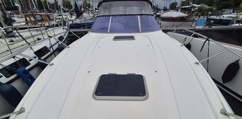 De foto toont het voordek van een witte boot met twee luiken en een voorruit, omringd door andere boten in een haven.