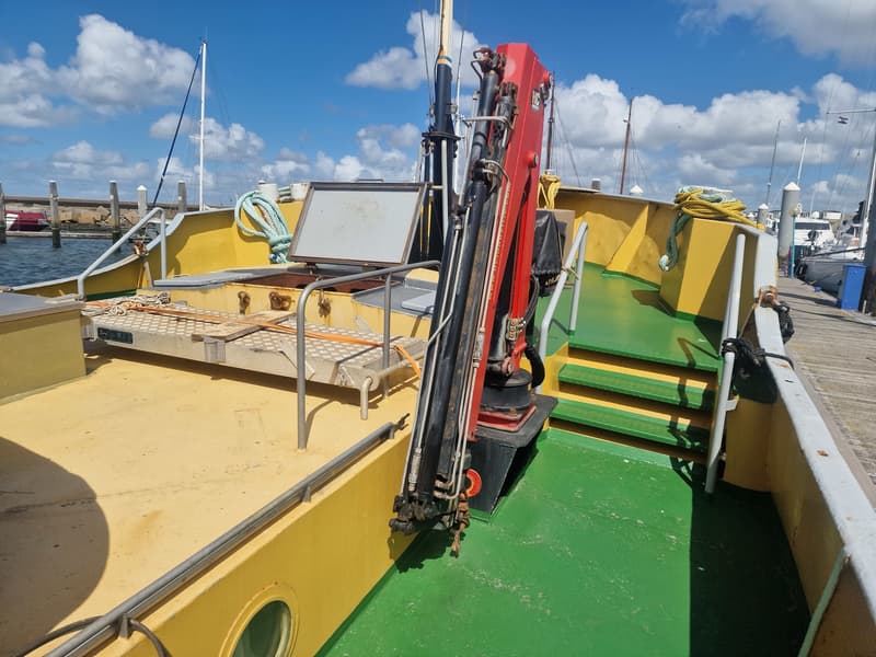 Een boot met een geel dek en groene vloer, een rode kraan, en een open luik, afgemeerd aan een houten steiger.