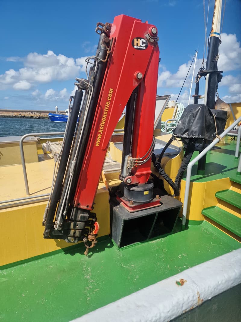 Een rode HC Industrie kraan staat op het groene dek van een boot, met een blauwe boot en de zee op de achtergrond.