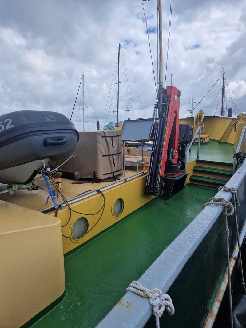 Een dek van een boot met een rubberboot, een rode kraan, patrijspoorten en zeilboten op de achtergrond.