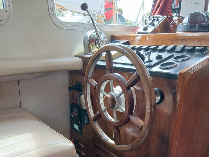 De foto toont een houten stuurwiel, een gashendel en diverse schakelaars en meters op een houten dashboard.