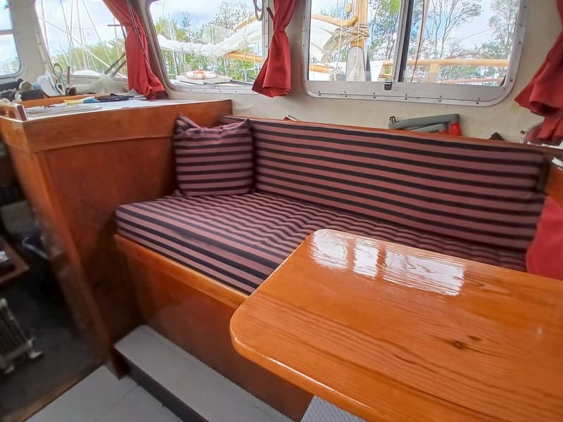 Een interieur van een boot met een houten tafel en een gestreepte bank met kussens, en ramen met rode gordijnen. — Fisher 34