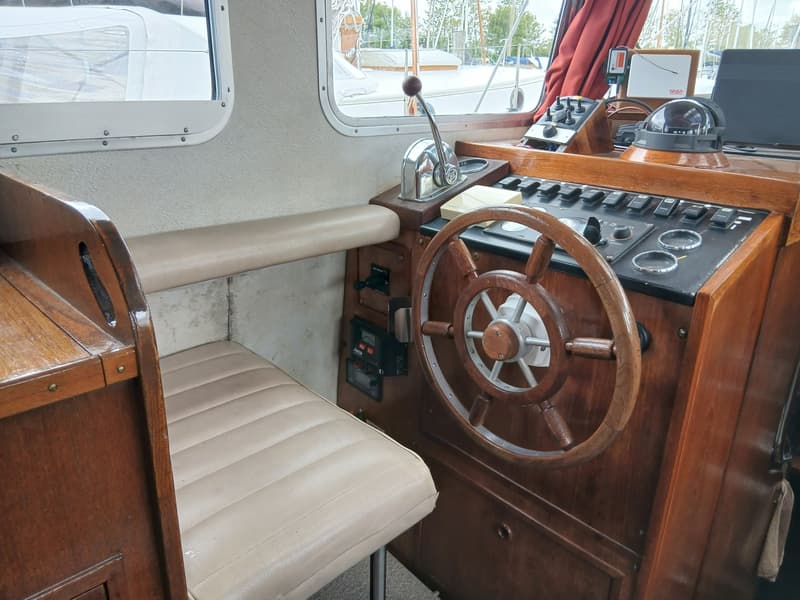 Een interieurfoto van een boot met een houten stuurwiel, een beige zitting en een dashboard met diverse bedieningselementen.