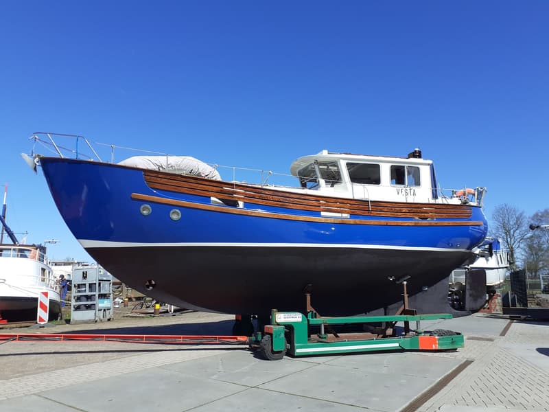 Een blauw-witte boot met houten accenten staat op een groene botenwagen op een betonnen ondergrond onder een helderblauwe…