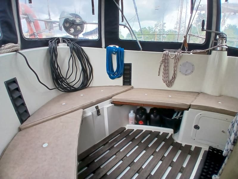 De foto toont een deel van een bootinterieur met beige kussens, touwen, een houten rooster op de vloer en ramen met…