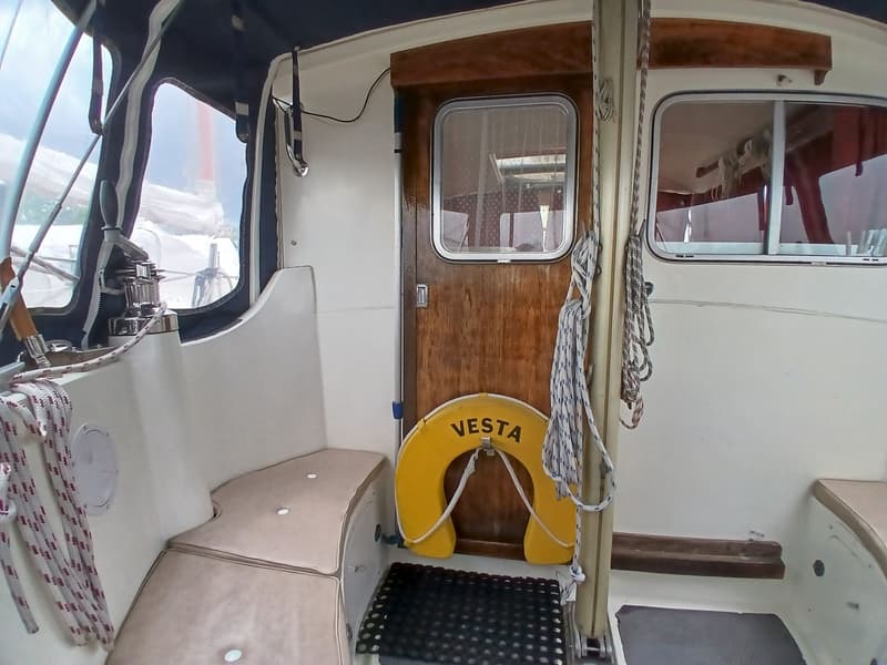 De foto toont een deel van een bootinterieur met een houten deur, ramen, touwen, een gele reddingsboei met "VESTA" erop en…