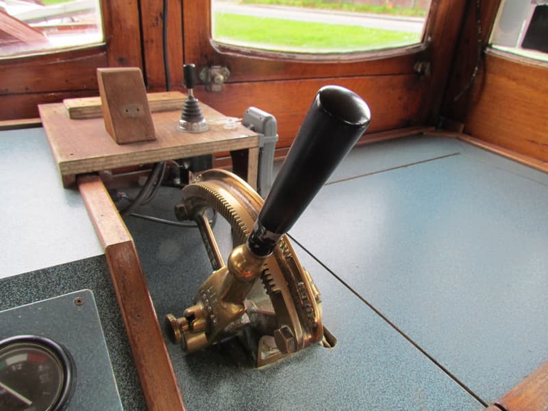 Een detailfoto van een bootinterieur met een koperen hendel, een houten plank en een raam met uitzicht op groen.
