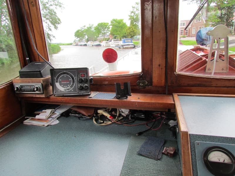 De foto toont het interieur van een stuurhuis met houten kozijnen en een uitzicht op een kanaal met aangemeerde boten.