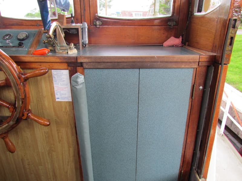 De foto toont een houten stuurwiel, een bedieningspaneel met meters en schakelaars, en twee blauwgrijze kastdeuren.