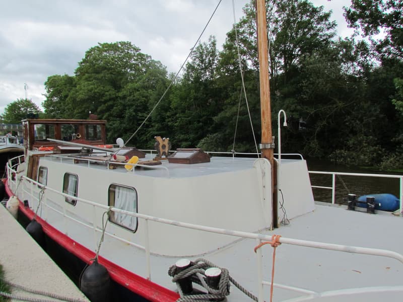 Een witte boot met rode en zwarte accenten ligt aangemeerd aan een kade, met een houten mast en stuurhut.