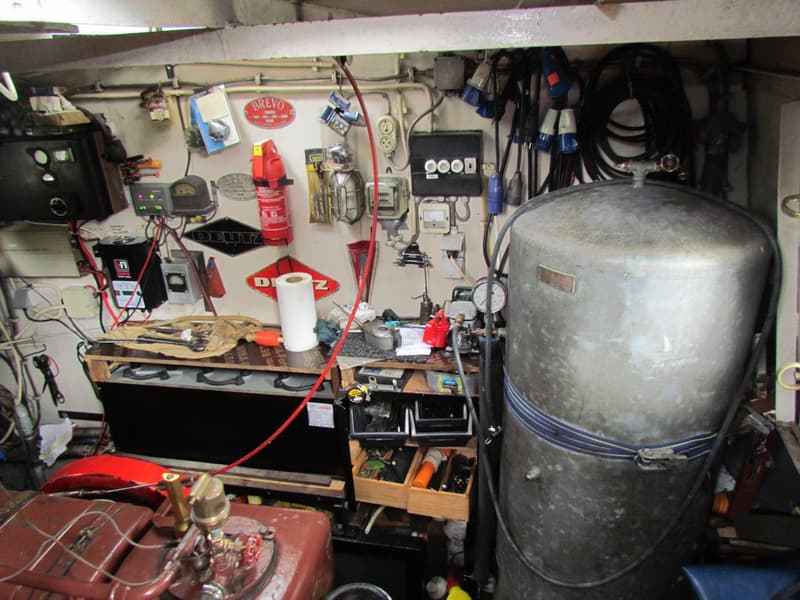 Een rommelige machinekamer met een grote grijze tank, diverse meters, kabels, gereedschap en een brandblusser.
