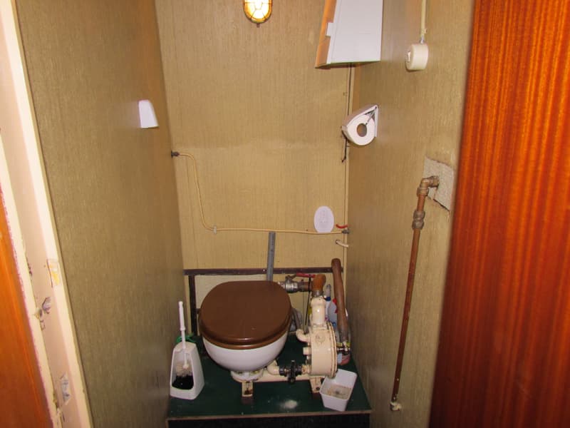 Een kleine toiletruimte met een wit toilet met bruine zitting, een toiletborstel, leidingen, een boiler en een lamp.