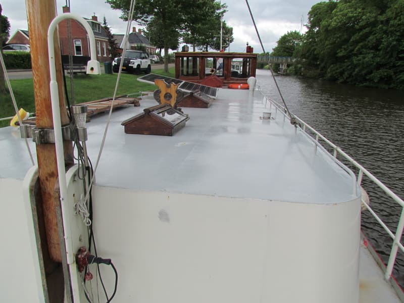 Een boot met een wit dek en reling vaart op een kanaal langs een groene oever met bomen en huizen.