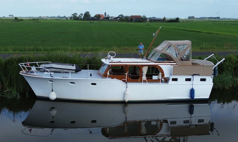 Een witte motorboot met houten details en een beige kap ligt in het water, met een man op de oever en een groen landschap…