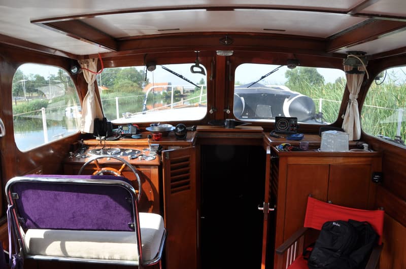Het interieur van een boot met houten afwerking, een stuurwiel, instrumentenpaneel, paarse stoel en uitzicht naar buiten.