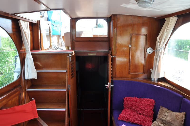 Een interieur van een boot met houten wanden, een trap, een paarse bank en een open deur naar een donkere ruimte.